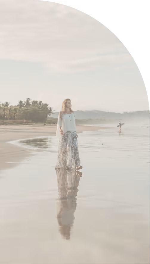 A woman in a white blouse and long skirt walking barefoot on a reflective wet sandy beach with a surfer carrying a surfboard in the background.