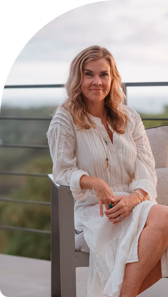 Smiling woman with wavy blonde hair wearing a white dress seated on a modern chair outdoors with a blurred scenic background.