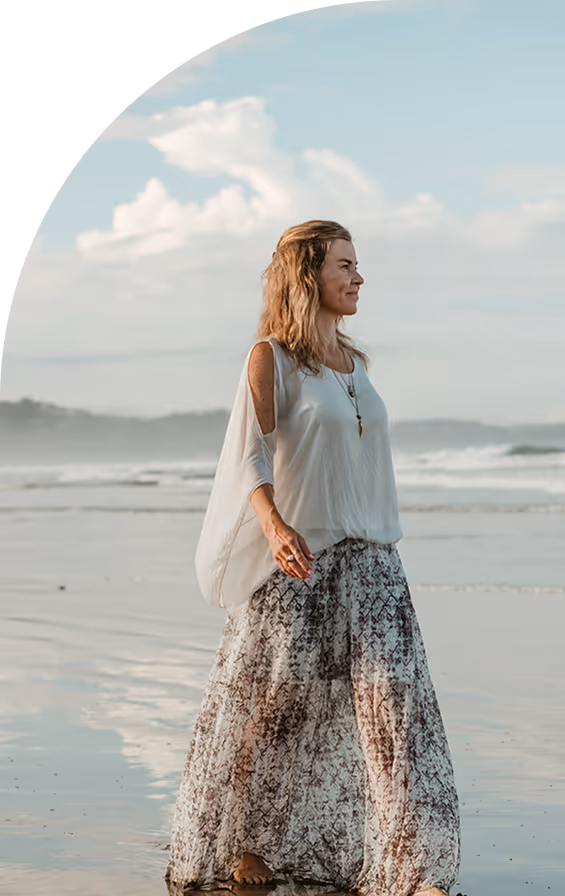 Woman in a white flowing top and patterned long skirt walking barefoot on a beach with waves and cloudy sky in the background.