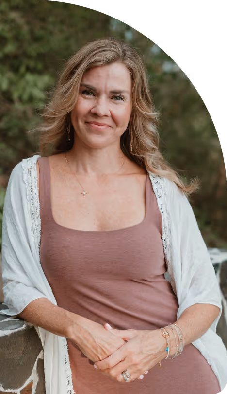 Smiling woman with wavy blonde hair wearing a mauve sleeveless dress and white shawl, seated outdoors.