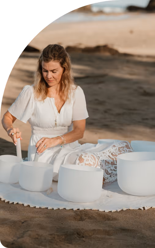 Woman in a white dress playing white crystal singing bowls on a sandy beach.
