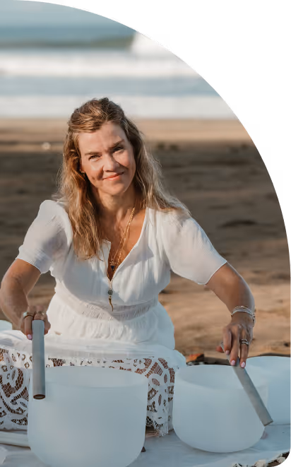 Smiling woman in white dress playing crystal singing bowls on a beach.