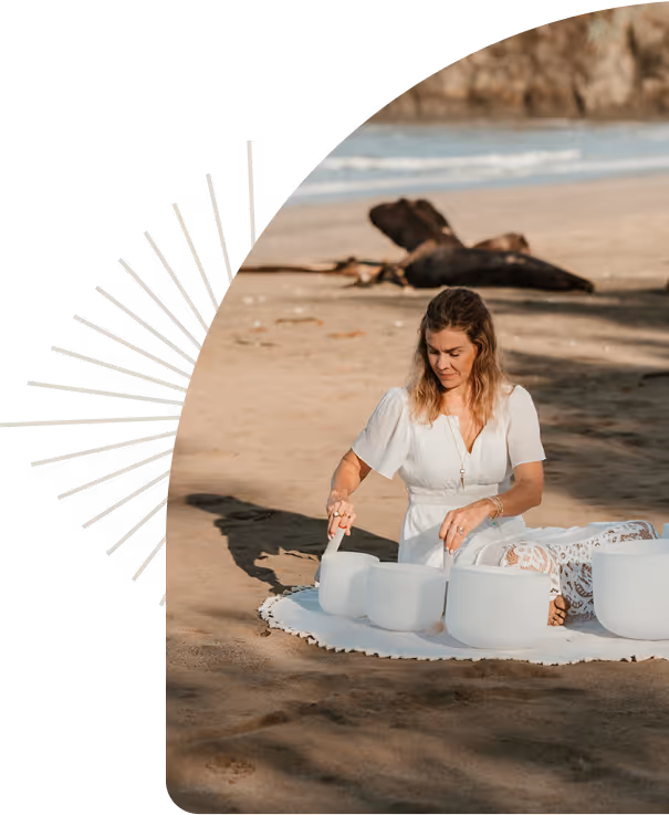Woman in white dress playing crystal singing bowls on a beach at sunset.