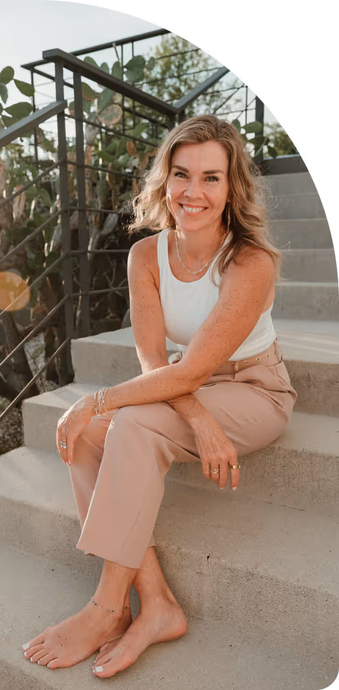 Smiling woman with wavy blonde hair, wearing a white sleeveless top and beige pants, sitting barefoot on outdoor concrete stairs.