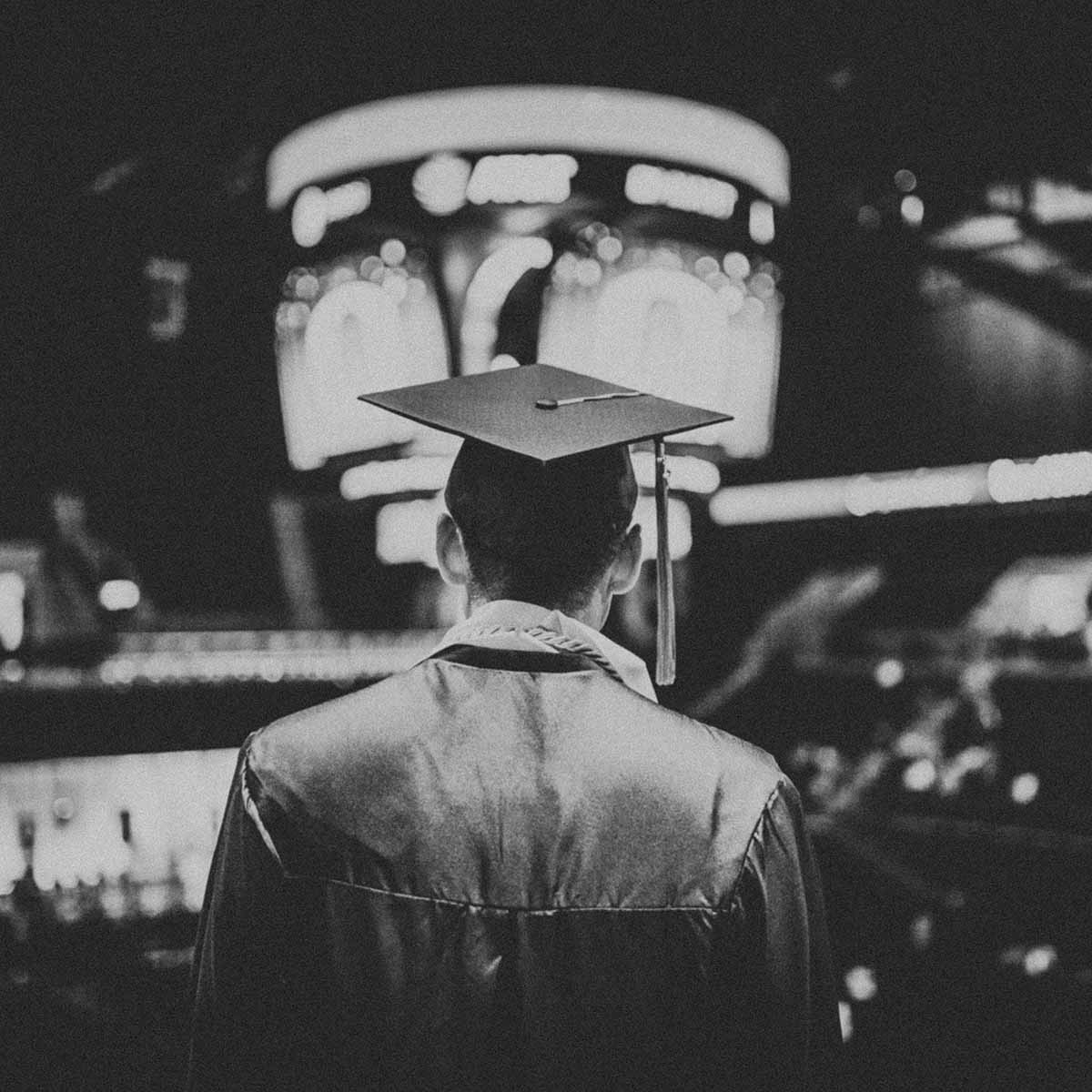 Graduate standing in auditorium, pictured from behind with cap on his head.