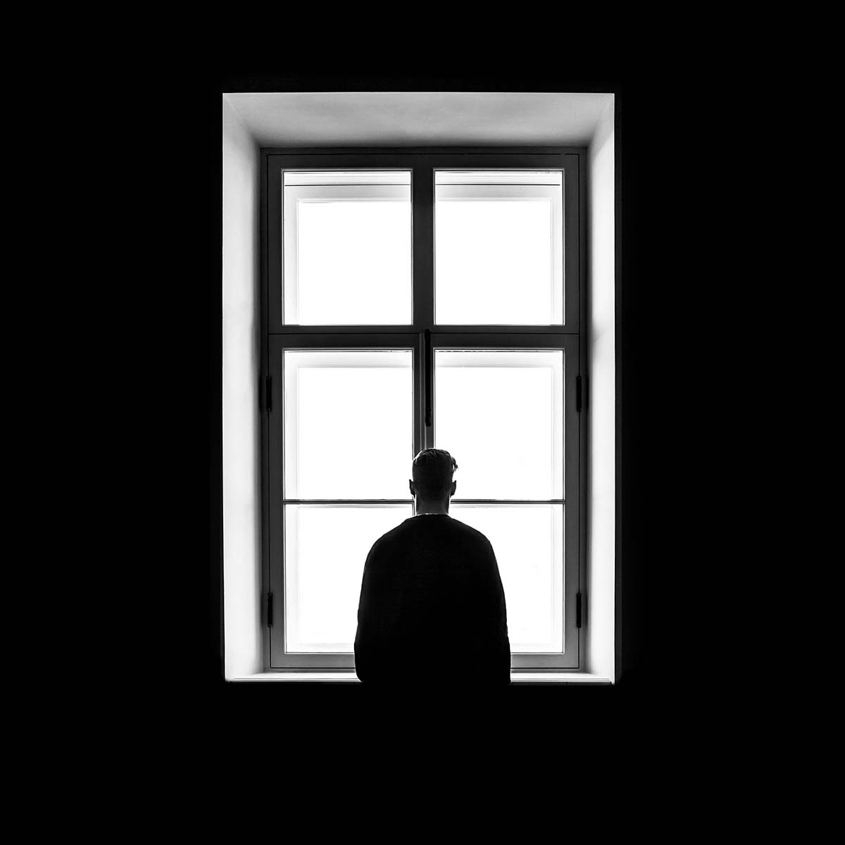 Black-and-white photo of a man looking out a tall window from within a dark room. Photo is taken from 15-20 feet behind the man, viewing his back.