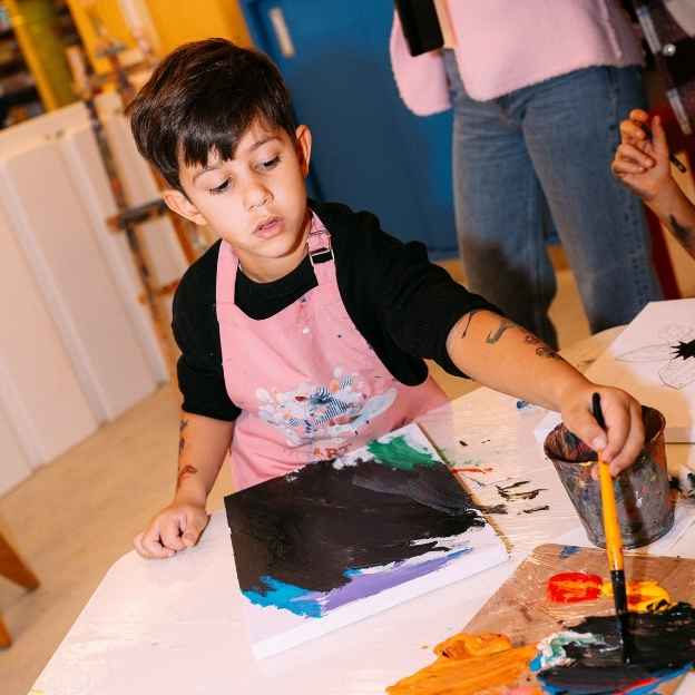 Young child in pink apron painting on paper with brush at art table indoors.