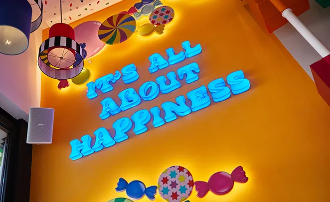 Bright blue neon sign reading "It's All About Happiness" on orange wall with colorful decorations.