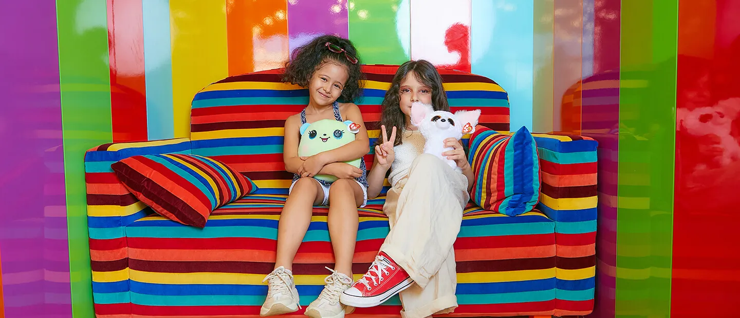 Two young girls sitting on colorful rainbow striped couch holding stuffed animals.