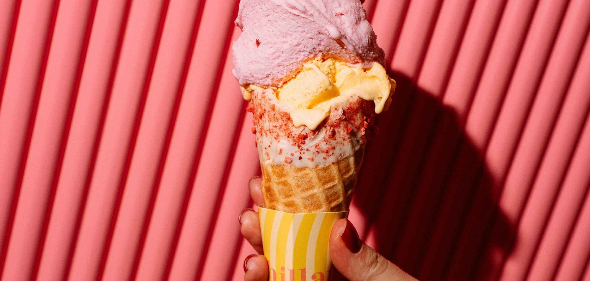 Hand holding colorful ice cream cone with pink, yellow and strawberry flavors against pink background