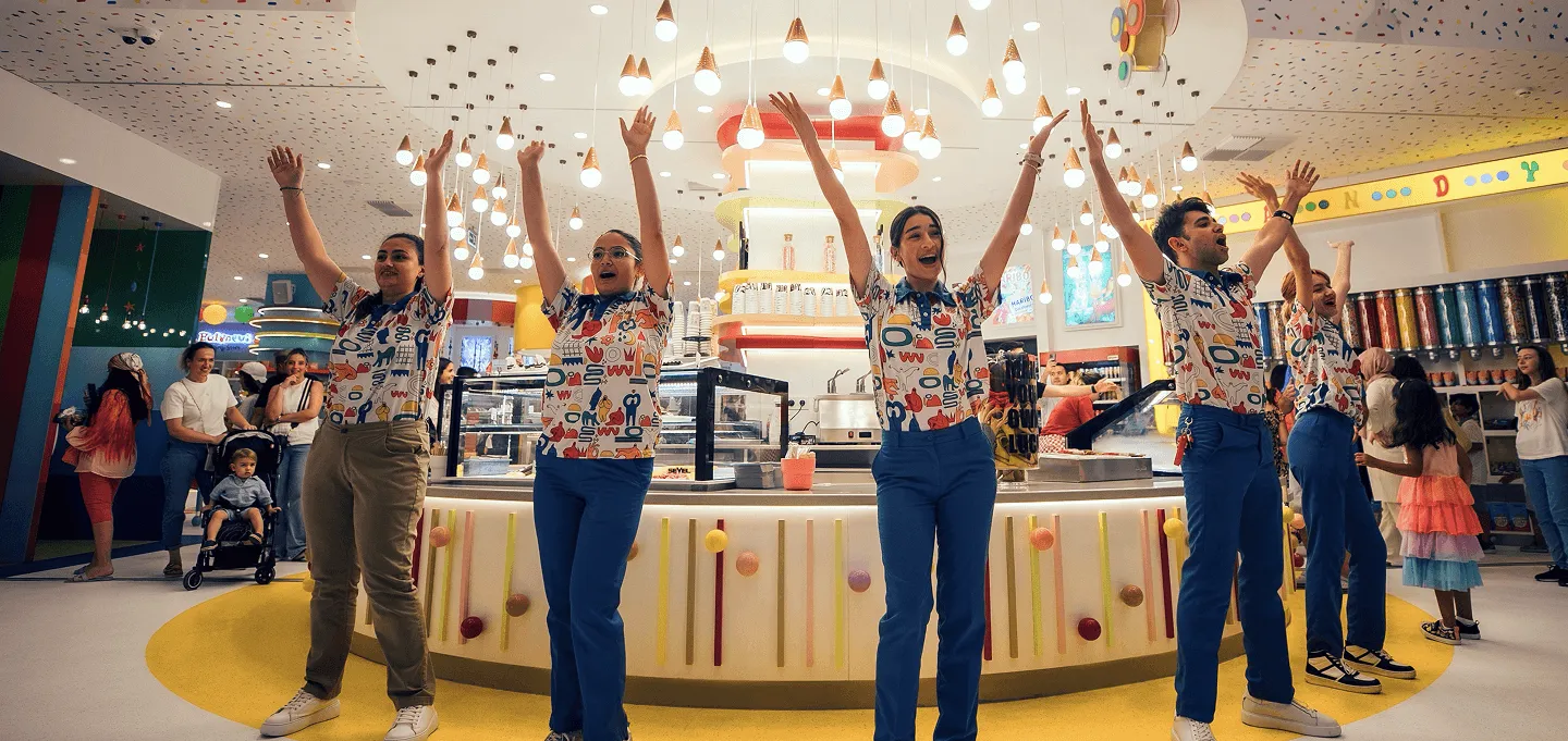 Staff members with raised arms celebrate in colorful indoor store with hanging decorations and shoppers.