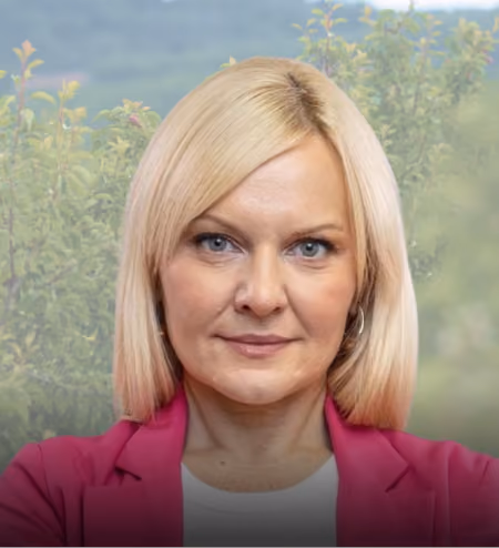 Woman with blonde hair wearing a pink blazer and white shirt, standing outdoors with greenery behind her.