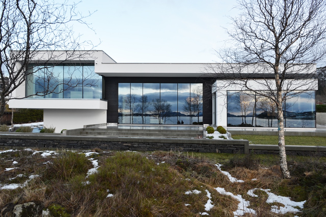 Fasadebilde av enebolig med panoramavinduer, flatt tak og speilende sjøutsikt i glassfeltene, omgitt av vinterlig vegetasjon