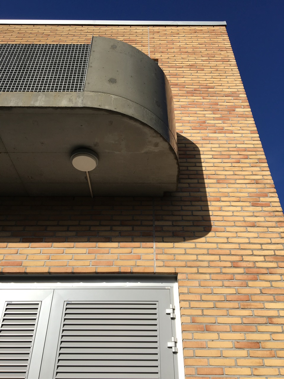 Detalj fra fasade med betongbalkong og buet kant, stålgitter og teknisk dør på gul teglbakgrunn.