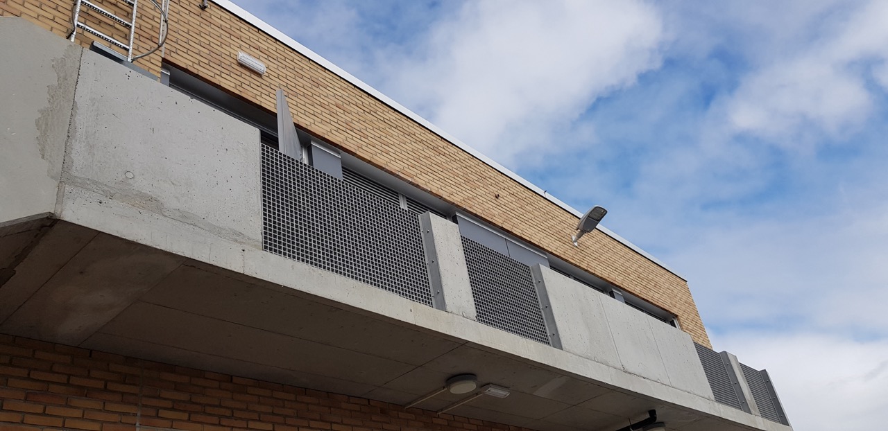 Balkong med betong og metallgitter, vinduer og tekniske installasjoner på oversiden av trafoanlegget.