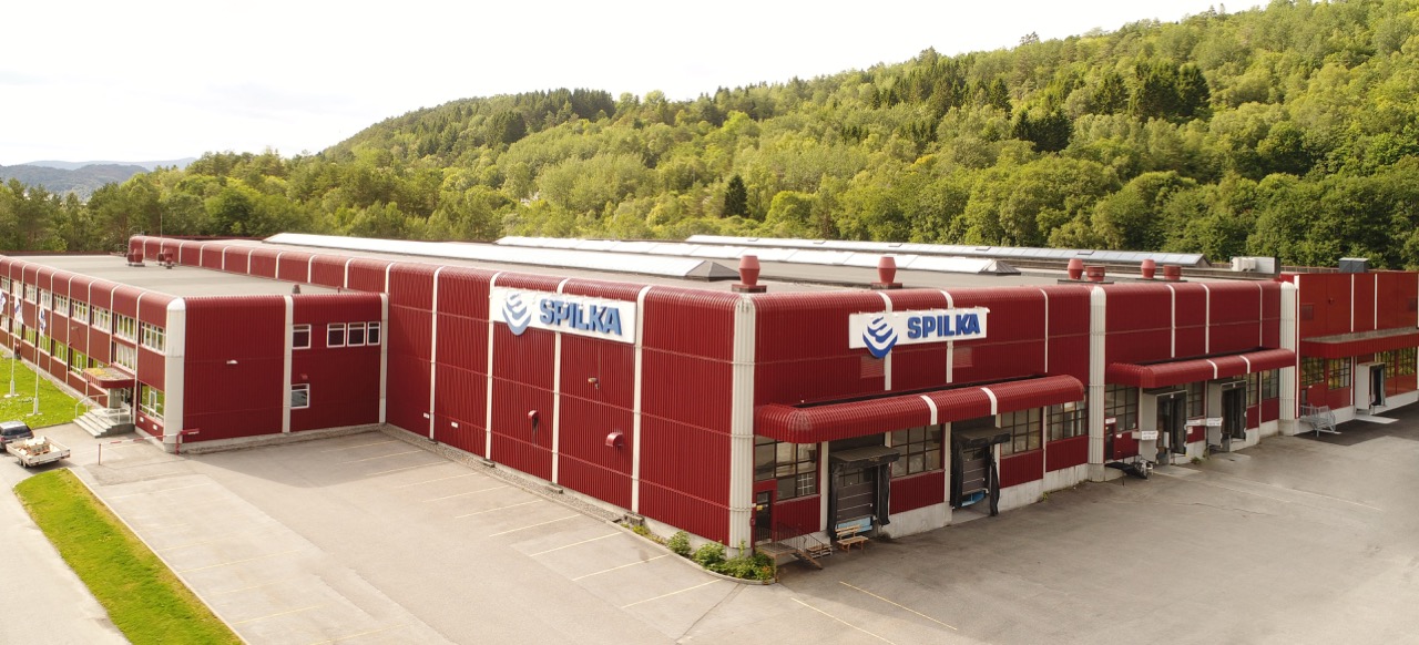 Fasadebilde av rødt industribygg med lasteporter, kontorfløy og Spilka-logo, omgitt av skog og asfaltflater.