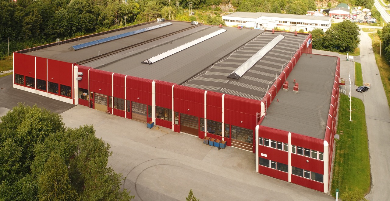 Luftfoto av industrianlegg med store produksjonshaller og takvinduer, omgitt av skogkledde områder.