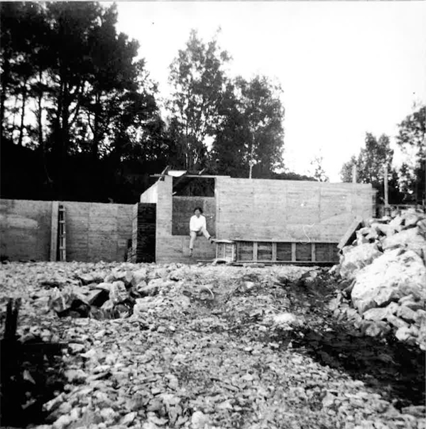 Sort-hvitt-bilde av støpearbeid og grunnmur under oppføring i kupert terreng med skog i bakgrunnen.