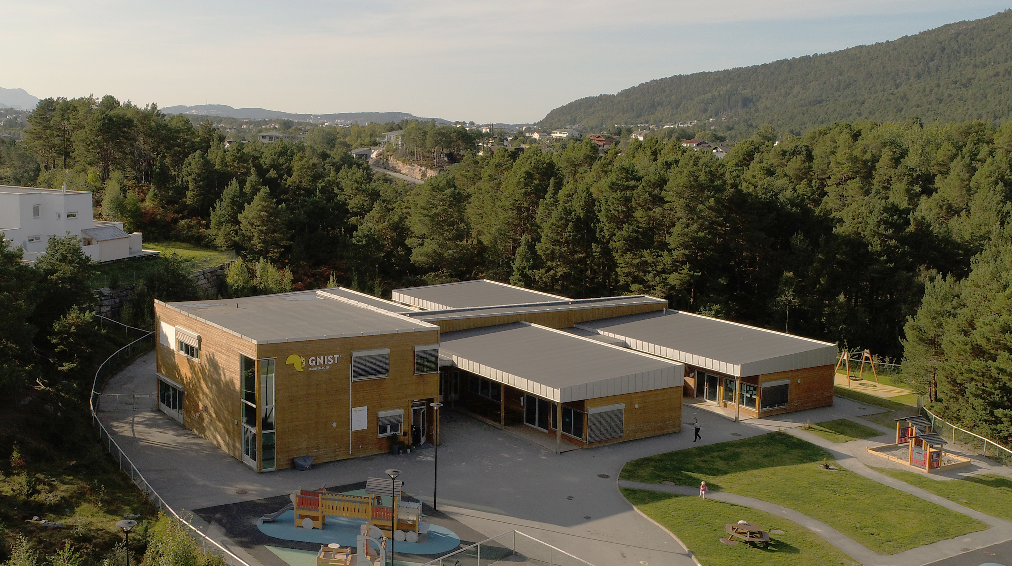 Dronefoto av Gnist-barnehage omgitt av skog og boligområde, med utendørs lekeplass og moderne trebygning.