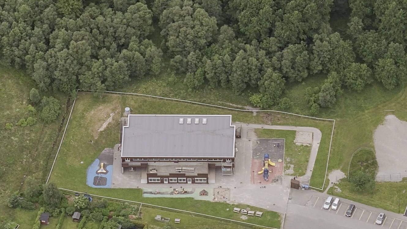 Loddrett dronefoto av barnehageanlegg med lekeplasser, asfaltflater og tilgrensende grøntområde.