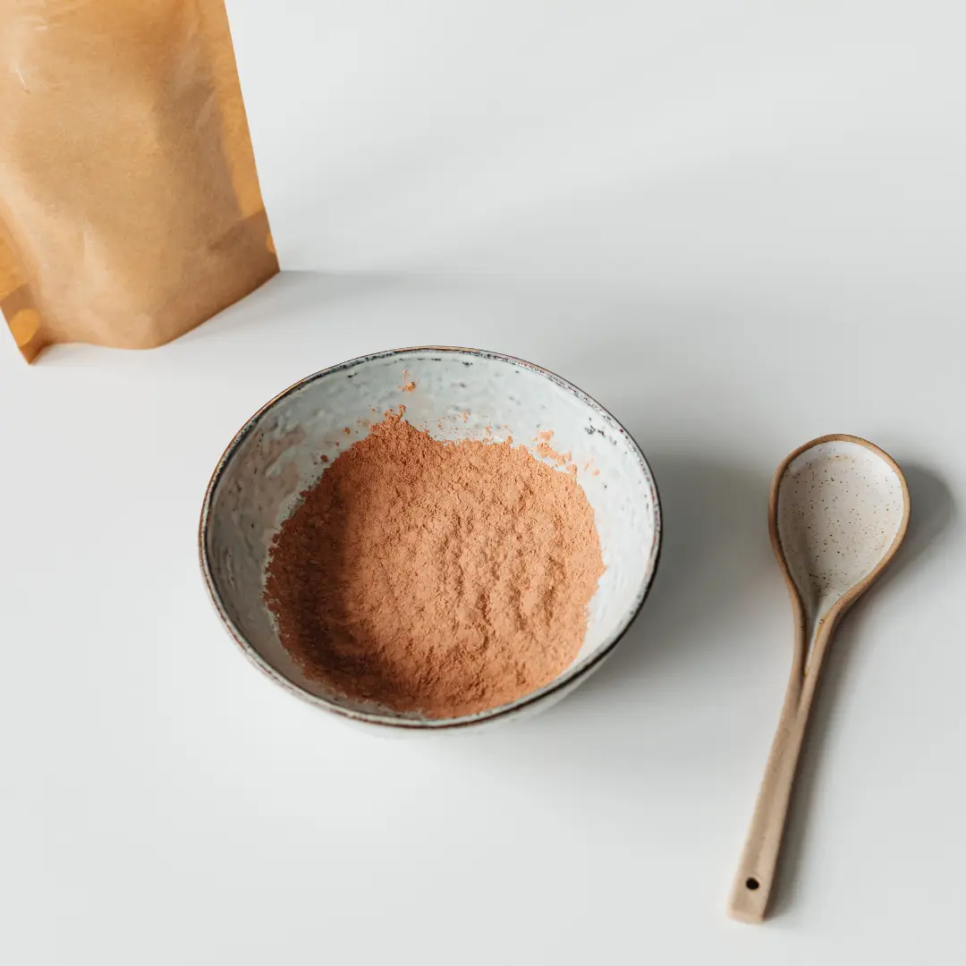 Bol en céramique contenant de la poudre de maca brun clair sur une surface blanche, à côté d'une cuillère en céramique et d'un sac en papier brun.
