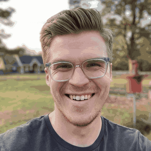 Smiling man with light brown hair and clear glasses outdoors with trees and houses in the background.