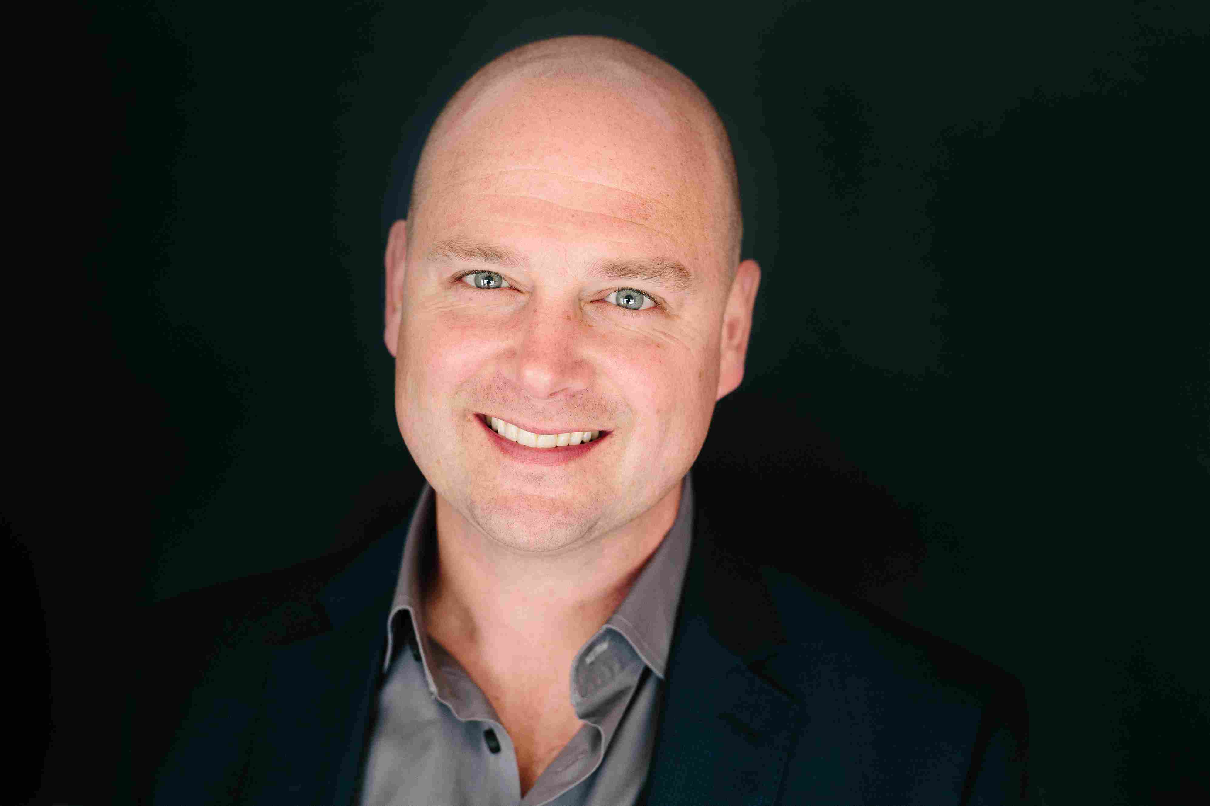 Smiling bald man wearing a dark blazer and gray shirt against a black background.