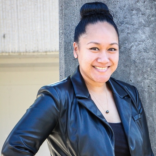 Smiling woman with dark hair in a bun wearing a black leather jacket and necklace, standing against a concrete wall.