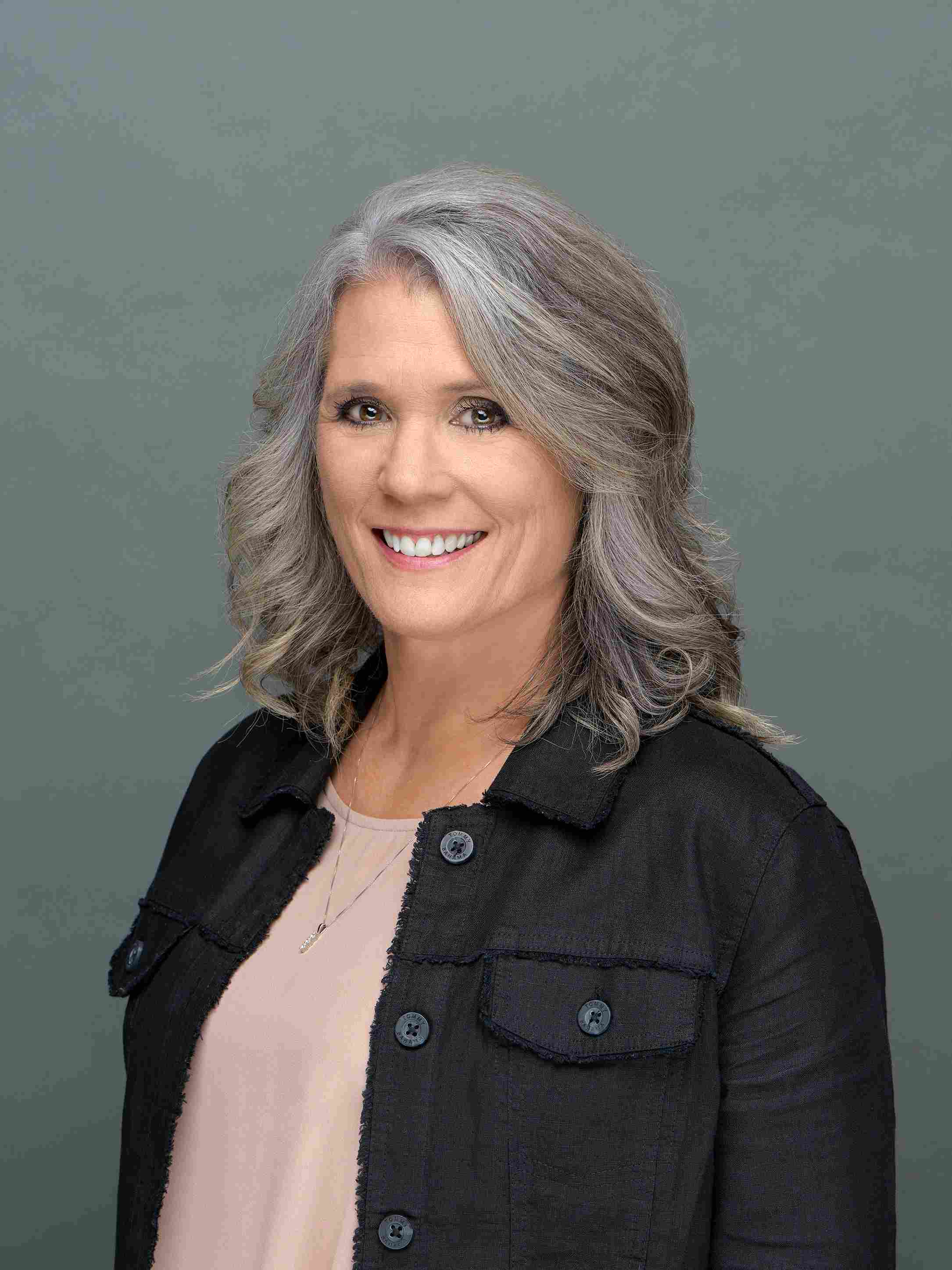Smiling middle-aged woman with shoulder-length gray hair wearing a black jacket over a light pink top against a gray background.