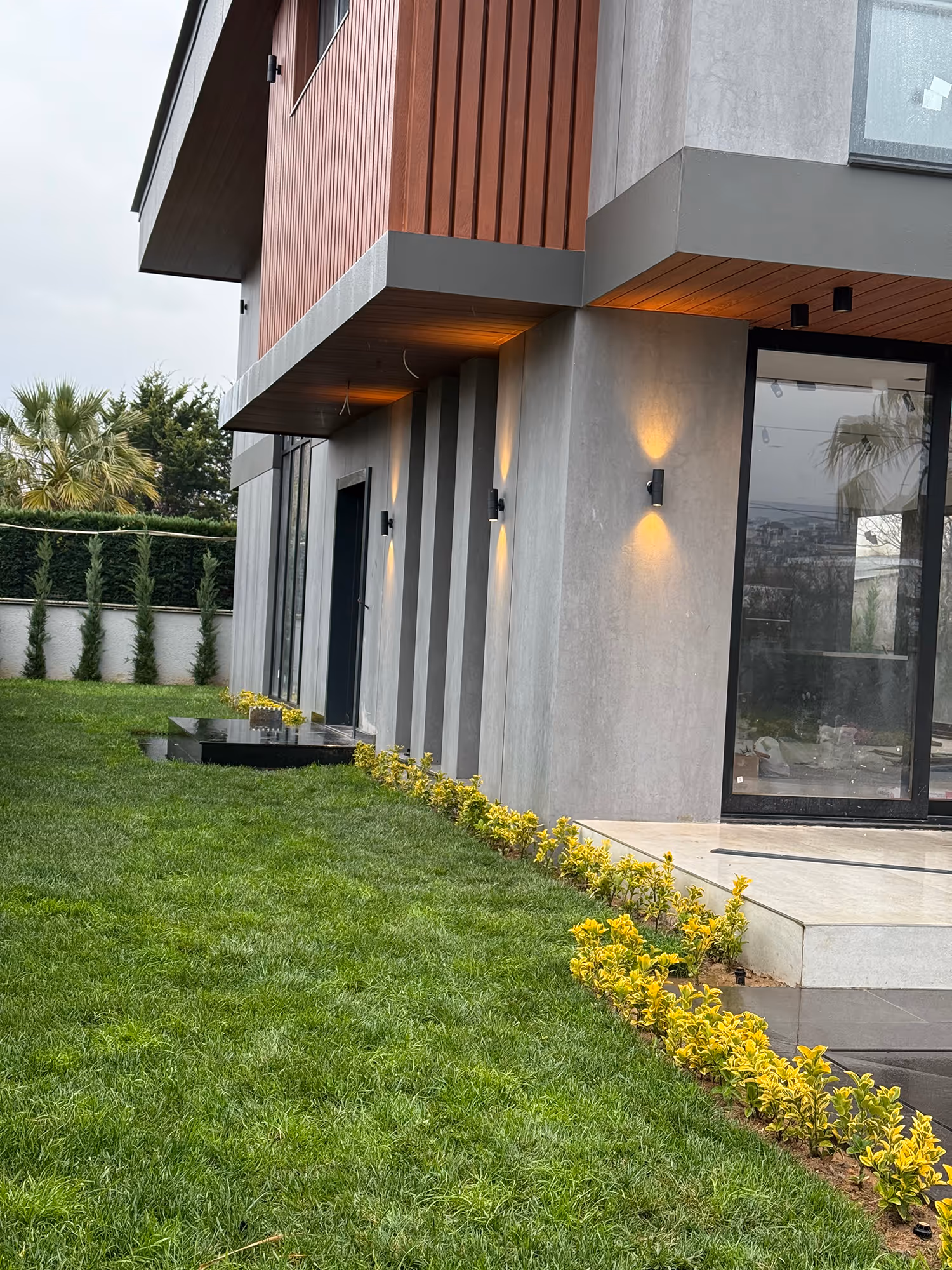Close-up view of modern villa exterior with concrete and wood paneling, accent wall lights, glass doors, manicured lawn, and neatly trimmed yellow shrubs along the foundation.