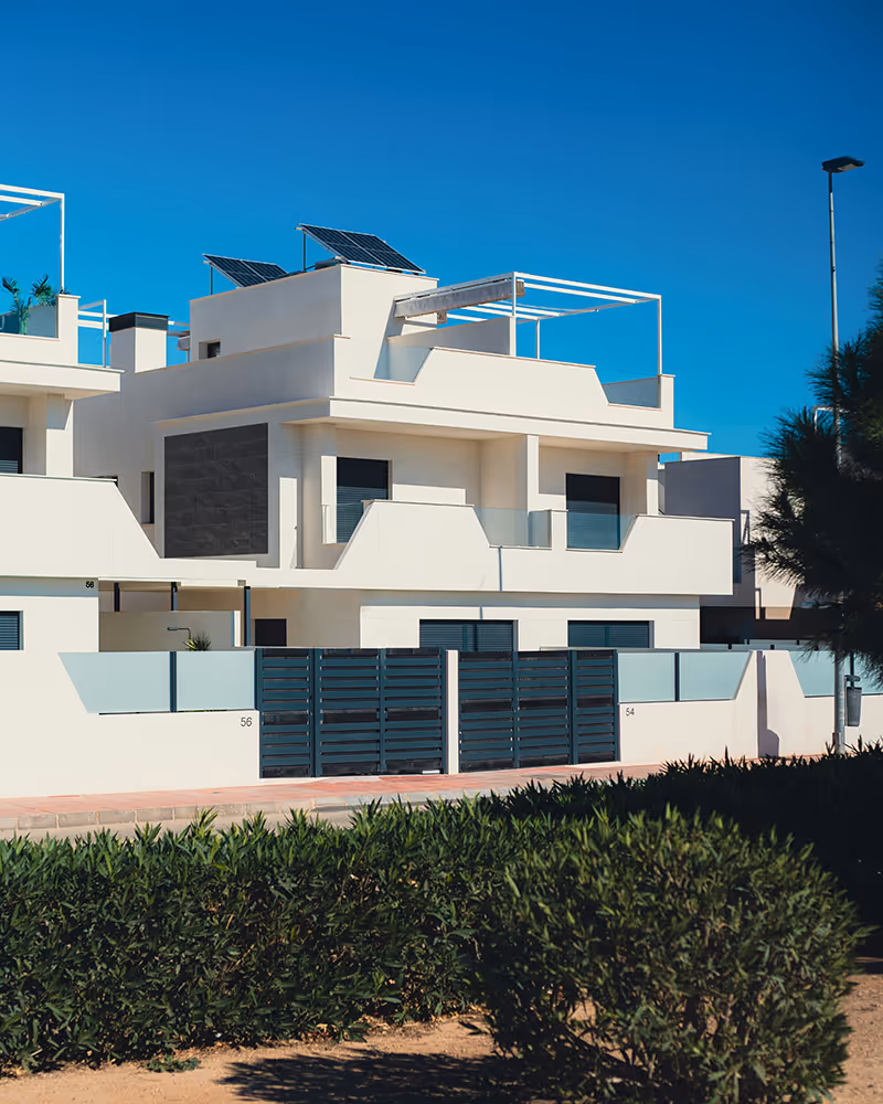 Modern white duplex with flat roofs, solar panels, glass balconies, and geometric design, surrounded by a black gate and shrubs, under clear blue sky in sunny residential neighborhood.