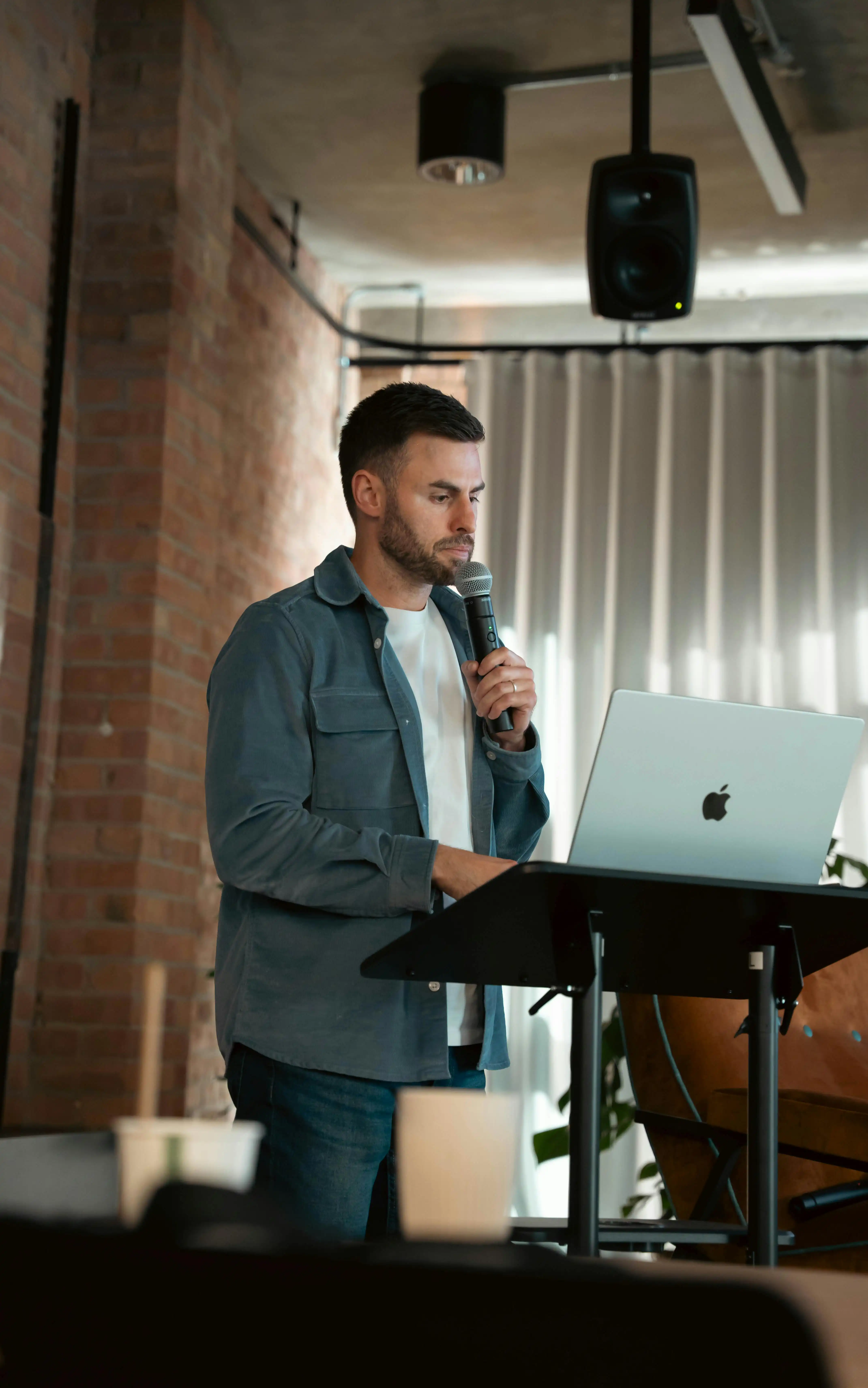 Man speaking into a microphone while standing at a podium with an open laptop in an indoor setting.