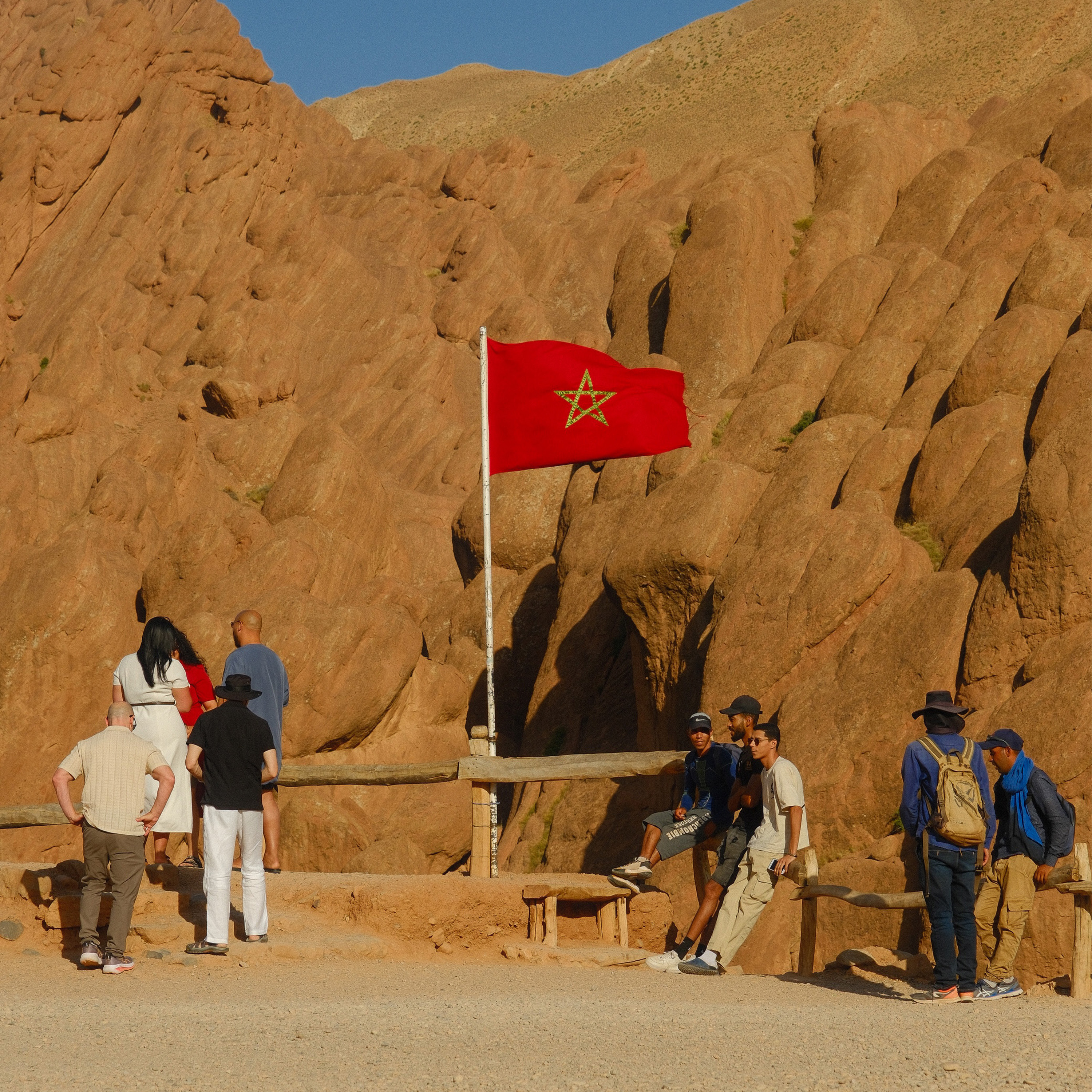 A man dressed in a vibrant orange robe and white scarf stands barefoot in the Moroccan desert, adjusting his head covering under a clear blue sky, surrounded by rolling dunes and golden sand.