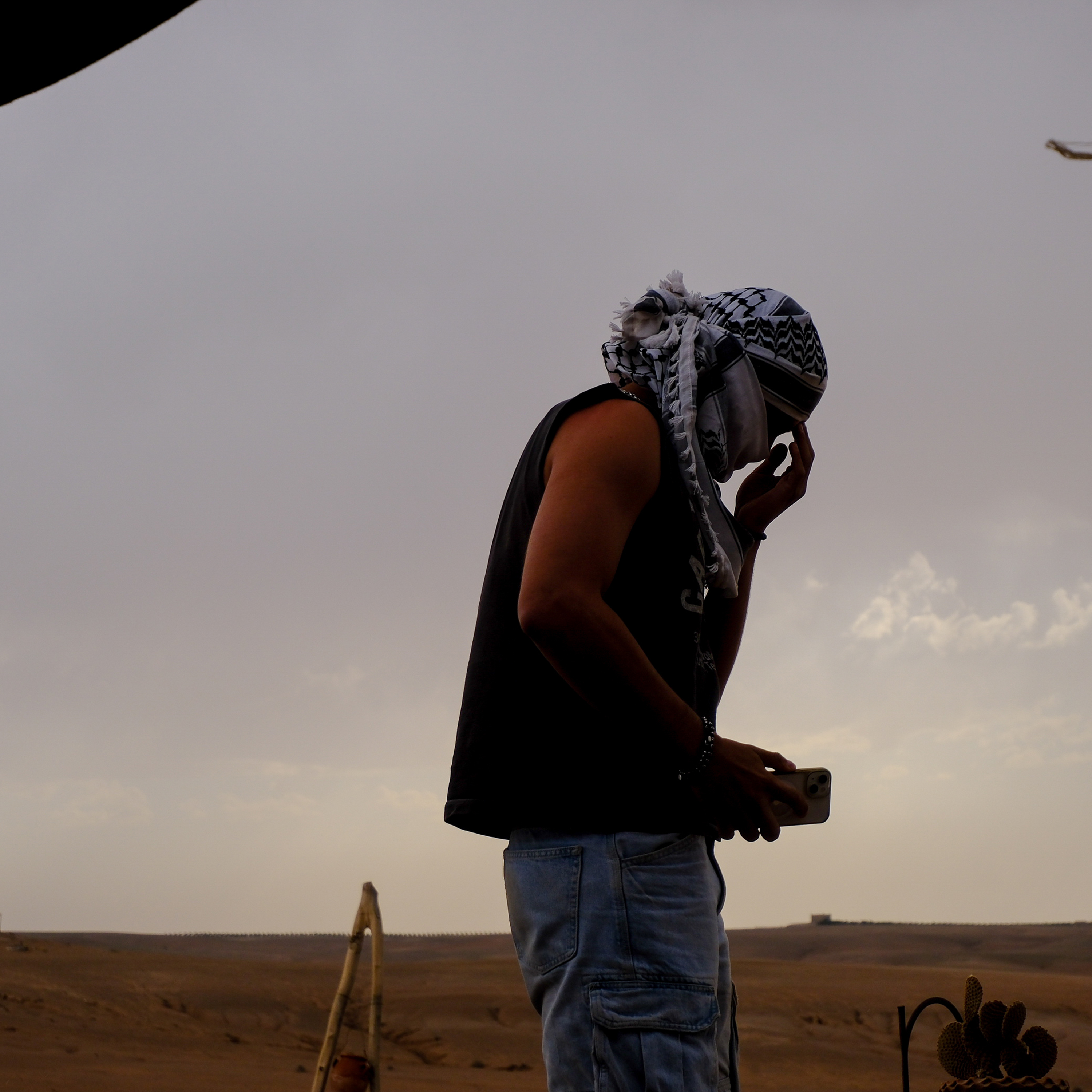 Silhouetted figure standing in a desert landscape at dusk, wearing a patterned headscarf and holding a smartphone, evoking themes of solitude and modern identity.
