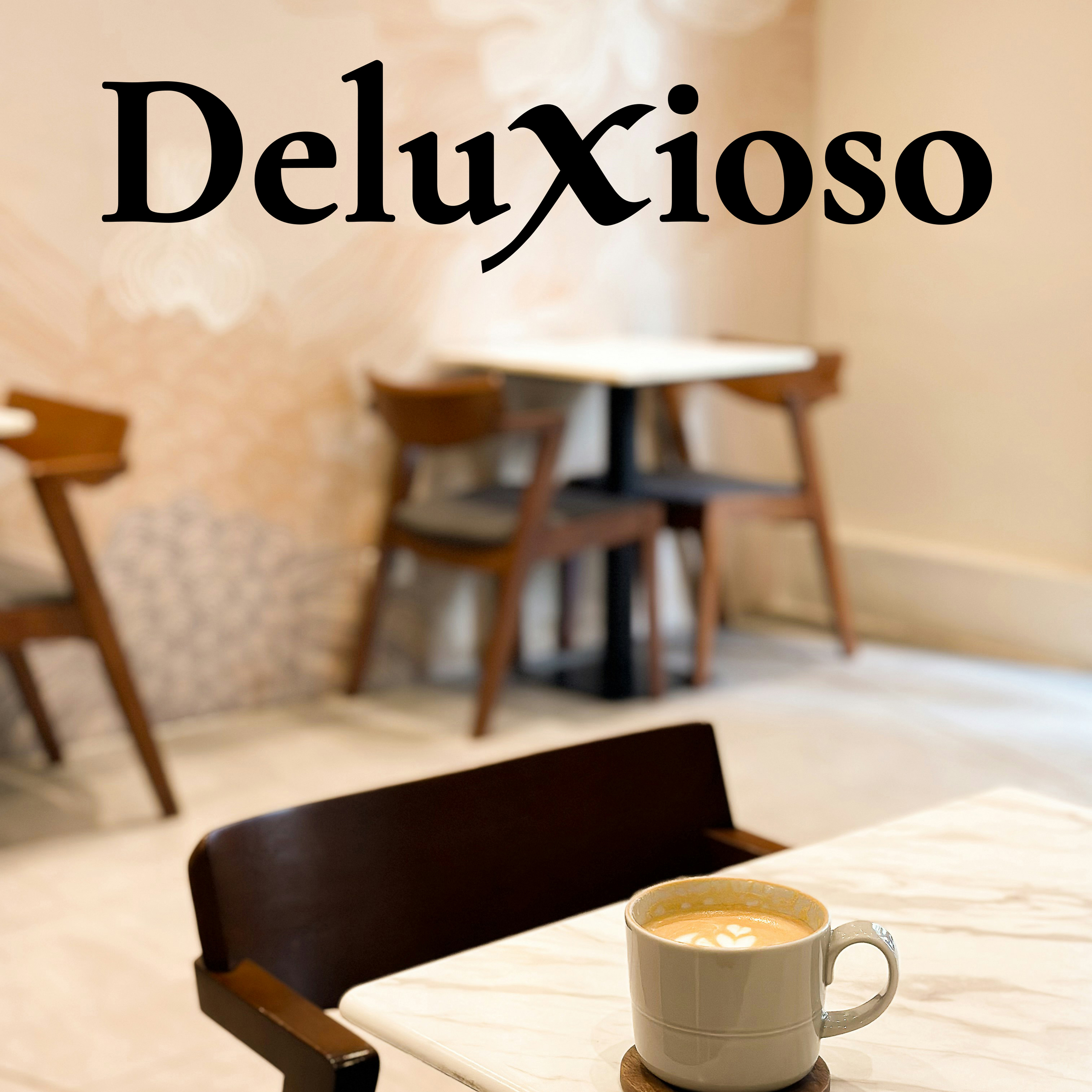 A ceramic coffee cup with latte art on a marble table in a softly lit café. The Deluxioso logo appears in black serif letters on the wall behind.