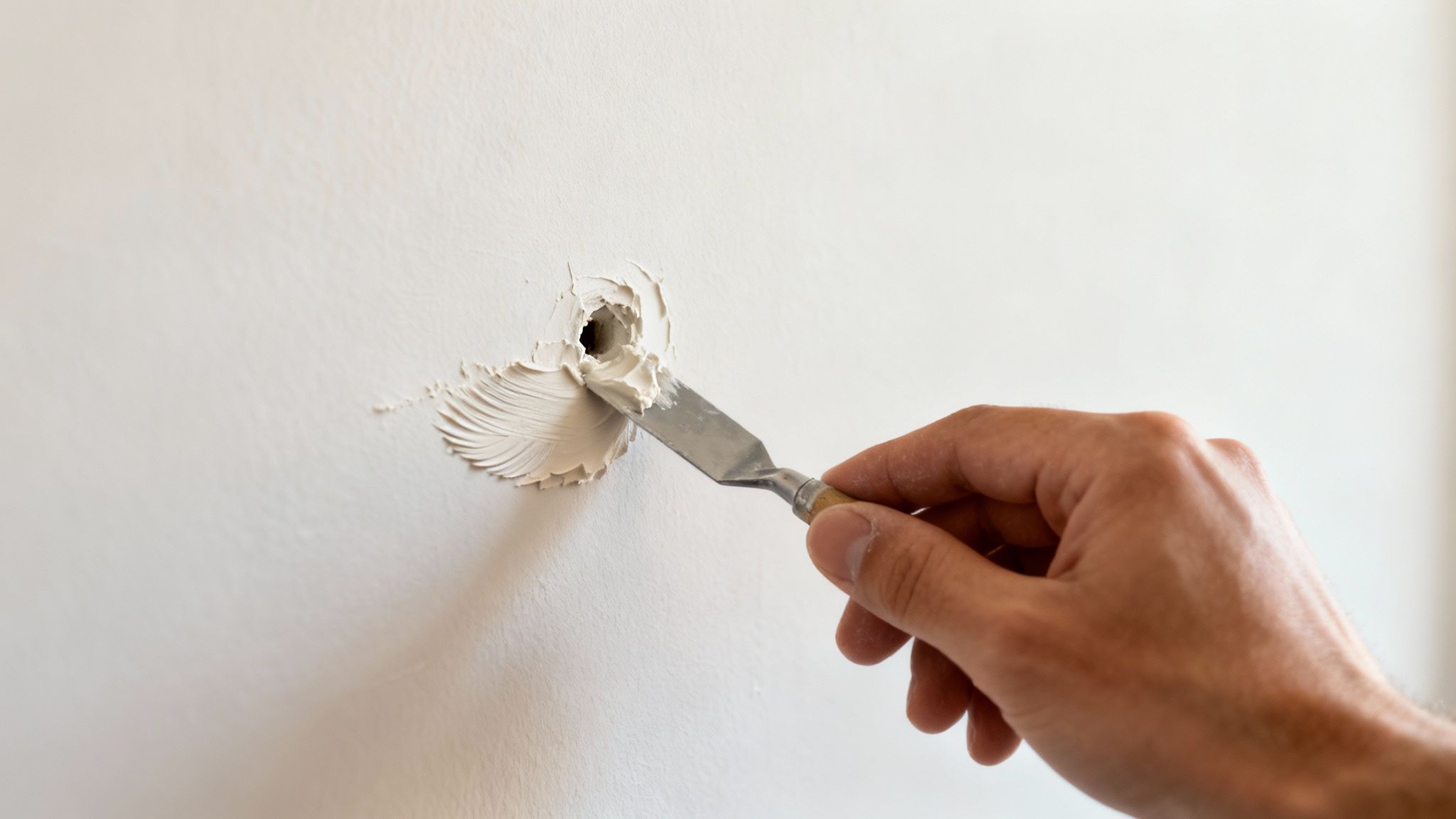 A hand expertly using a small spatula to apply white spackle, filling a hole in a clean white wall.