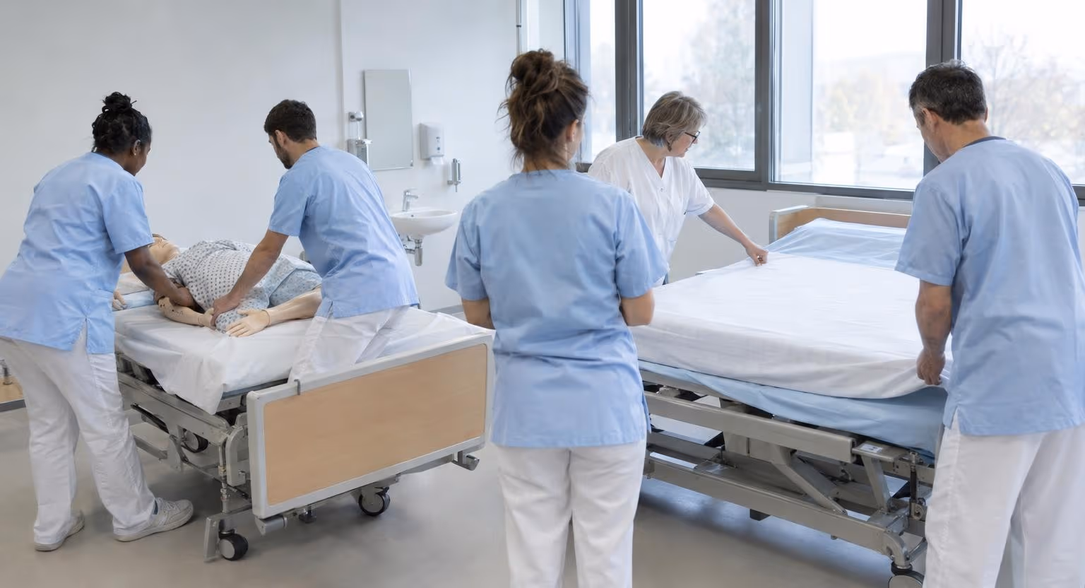Des adultes en tunique bleu pâle s’exercent à retourner un mannequin sur un lit médicalisé, pendant qu’une femme en blouse blanche montre un pli du drap.