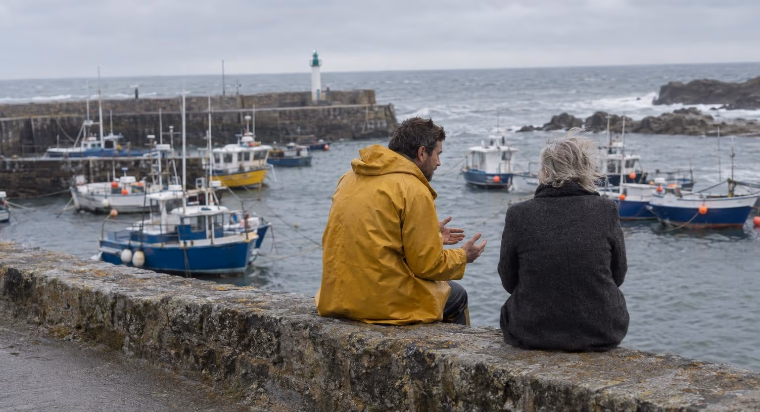 Un homme en ciré jaune et une femme âgée en manteau de laine, assis de dos sur le muret d’une jetée, discutent face au port et à la mer grise.