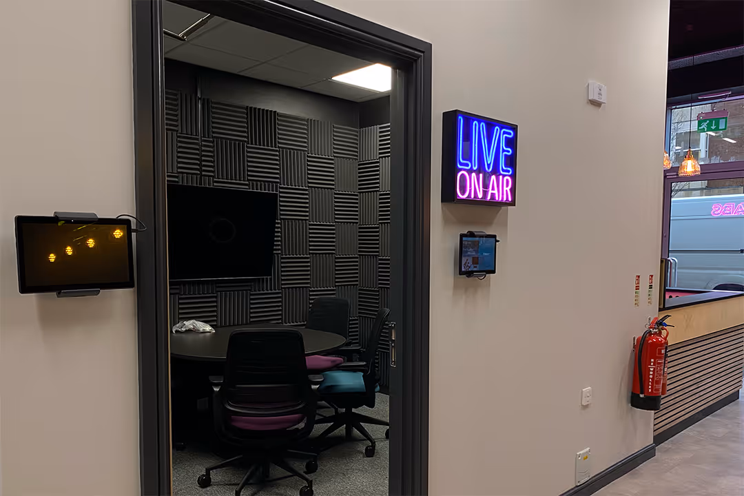 Recording studio room with foam soundproofing panels, round table with office chairs, and a neon sign reading LIVE ON AIR outside the door.