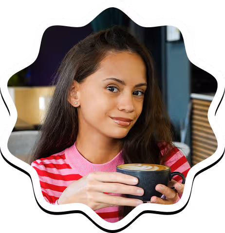 Woman with long dark hair wearing a pink and red striped shirt holding a cup of coffee with latte art in a cozy café.