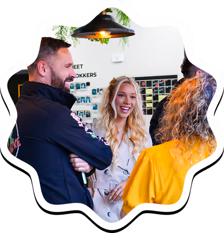 Three people smiling and chatting indoors under a hanging light with photos on the wall behind them.