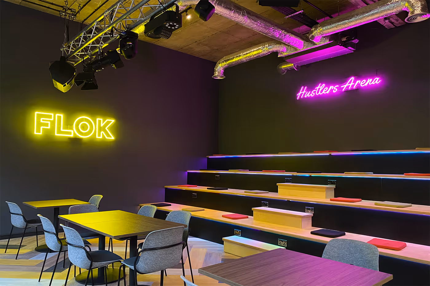 Modern event space with tiered seating, colorful cushions, grey chairs, and neon signs saying FLOK and Hustlers Arena.