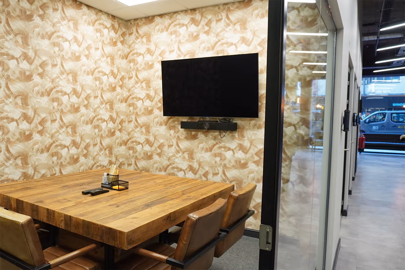 Small meeting room with a wooden table, four brown chairs, patterned wall, and a wall-mounted TV.