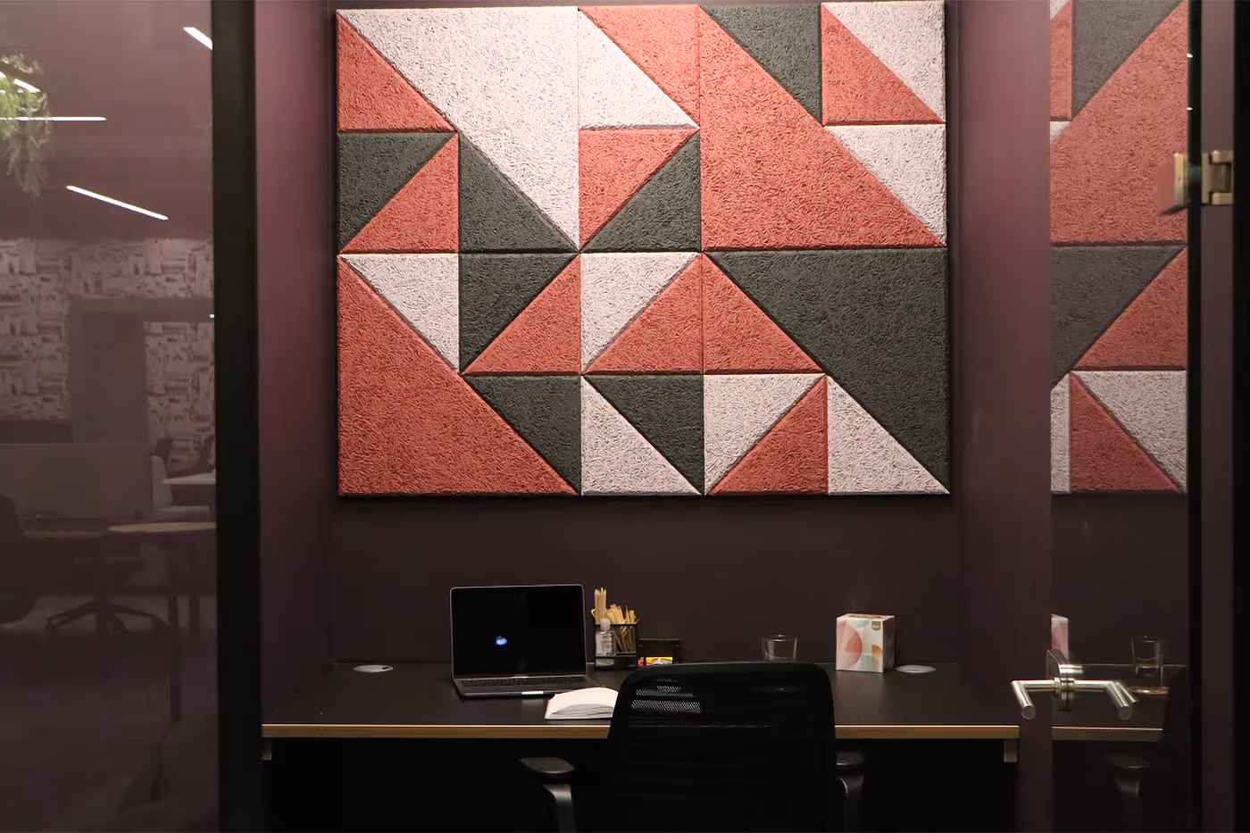 Small office space with a desk, laptop, chair, and wall art of red, pink, and green geometric shapes.