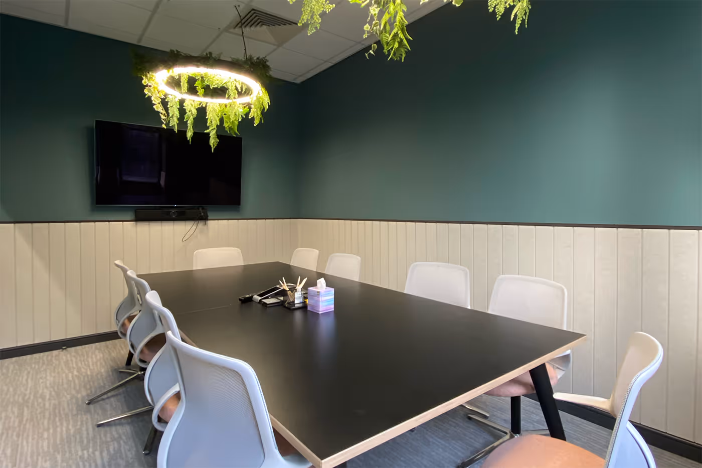 Modern conference room with black table, white chairs, green walls, TV, and hanging ring light with plants.