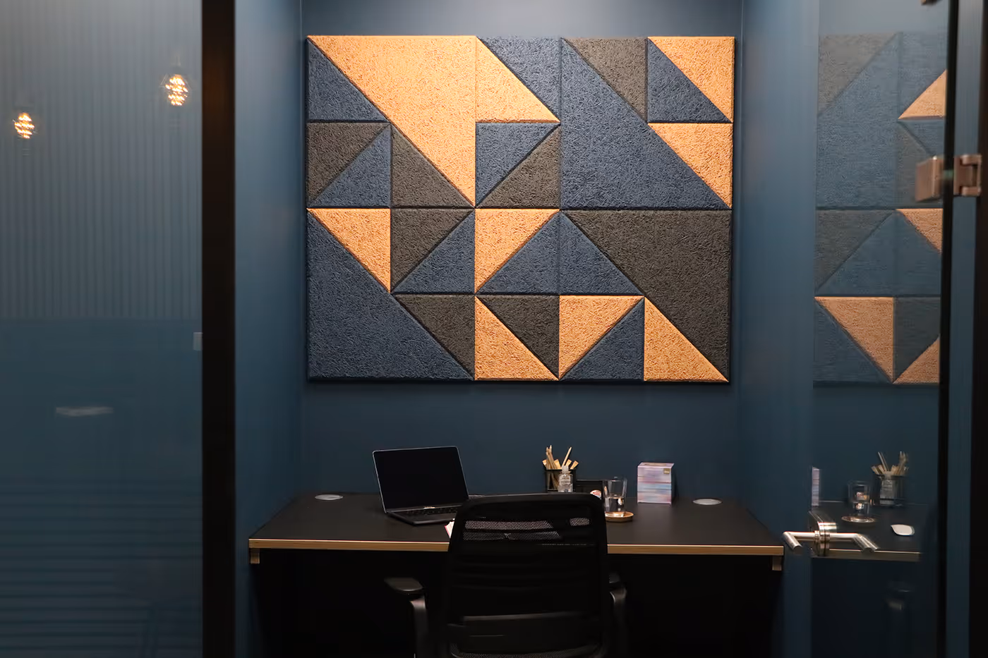 Small office with dark desk, black chair, laptop, and geometric wall art in blue and orange.