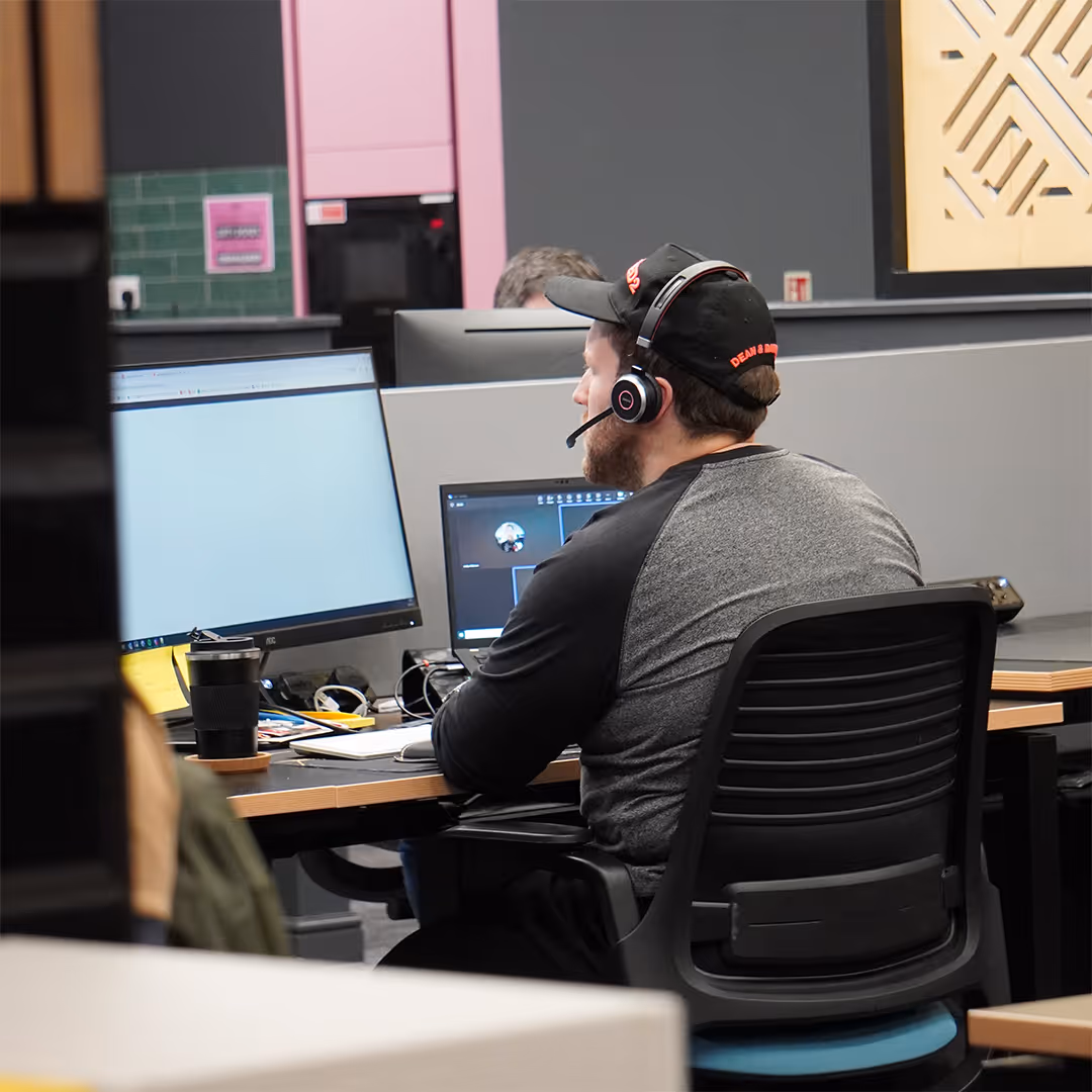 Man wearing a headset and cap working on a desktop computer with two monitors in an office setting.