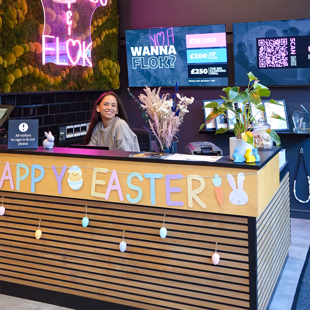Reception with smiling woman, Easter decorations, neon sign, and screens showing membership deals and QR code.