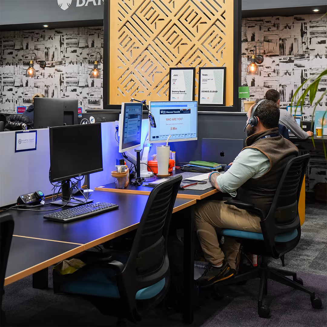Man wearing headset works at a desk with dual monitors in a modern office workspace.