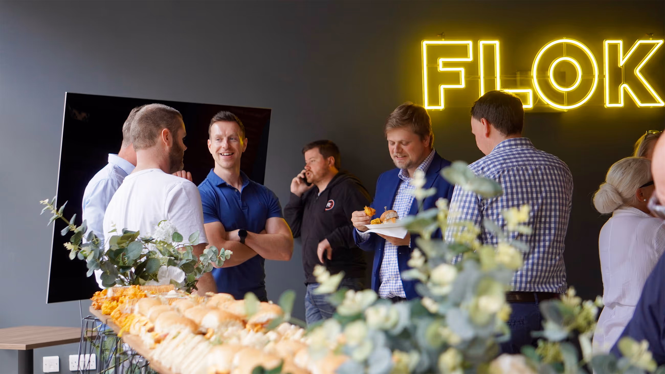 Group of people socializing near a table with food under a neon 'FLOK' sign on a gray wall.
